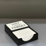 notepad with the words SH*T I MIGHT FORGET imprinted on a black acrylic holder with black letters, white plate displayed on a kitchen counter.